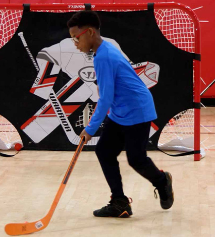little Black boy in blue top and black shorts using an orange hockey stick and playing with a little orange ball