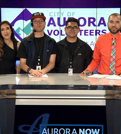 group of people standing behind the Aurora Now news desk