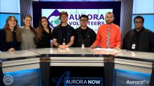 AuroraTV volunteers standing with AuroraTV anchors behind studio desk