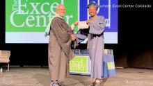 man in black graduation gown shaking hands and handing diploma to man in blue graduation gown