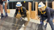 an African American man and blonde-haired woman wearing white hard hats and installing a solar panel