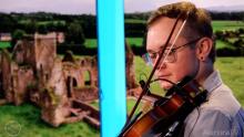 man playing the violin in front of a picture of a castle in a green Irish pasture