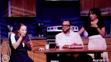 teen girl eating at table and seated angry man watching her along with stunned, standing woman