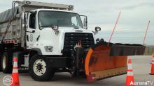close up image of a city of Aurora snowplow with a giant orange plow driving past orange traffic cones on dry surface